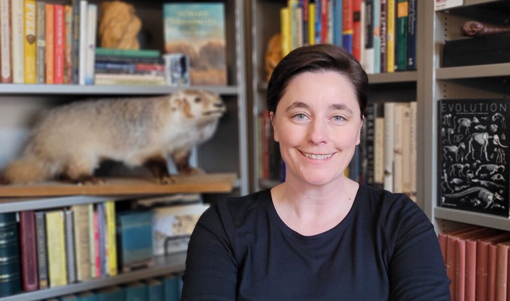 Smiling person in a book-lined office with taxidermy mammal display and evolution books on shelves.