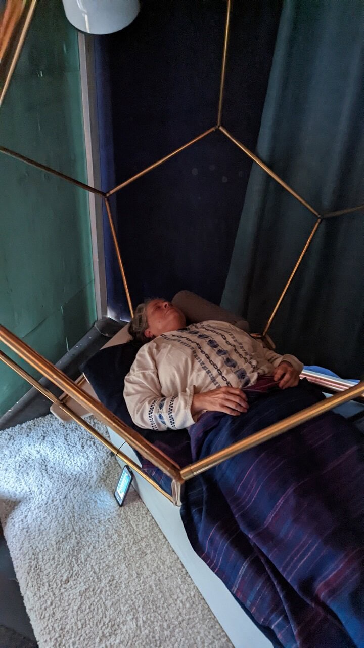 Person meditating or resting in a gold plated dodecahedron bed, wrapped in blanket in dim wellness room.
