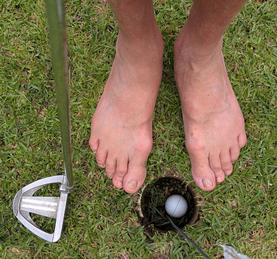 Bare feet on a golf green beside a putter and a ball sunk in the hole during putting practice