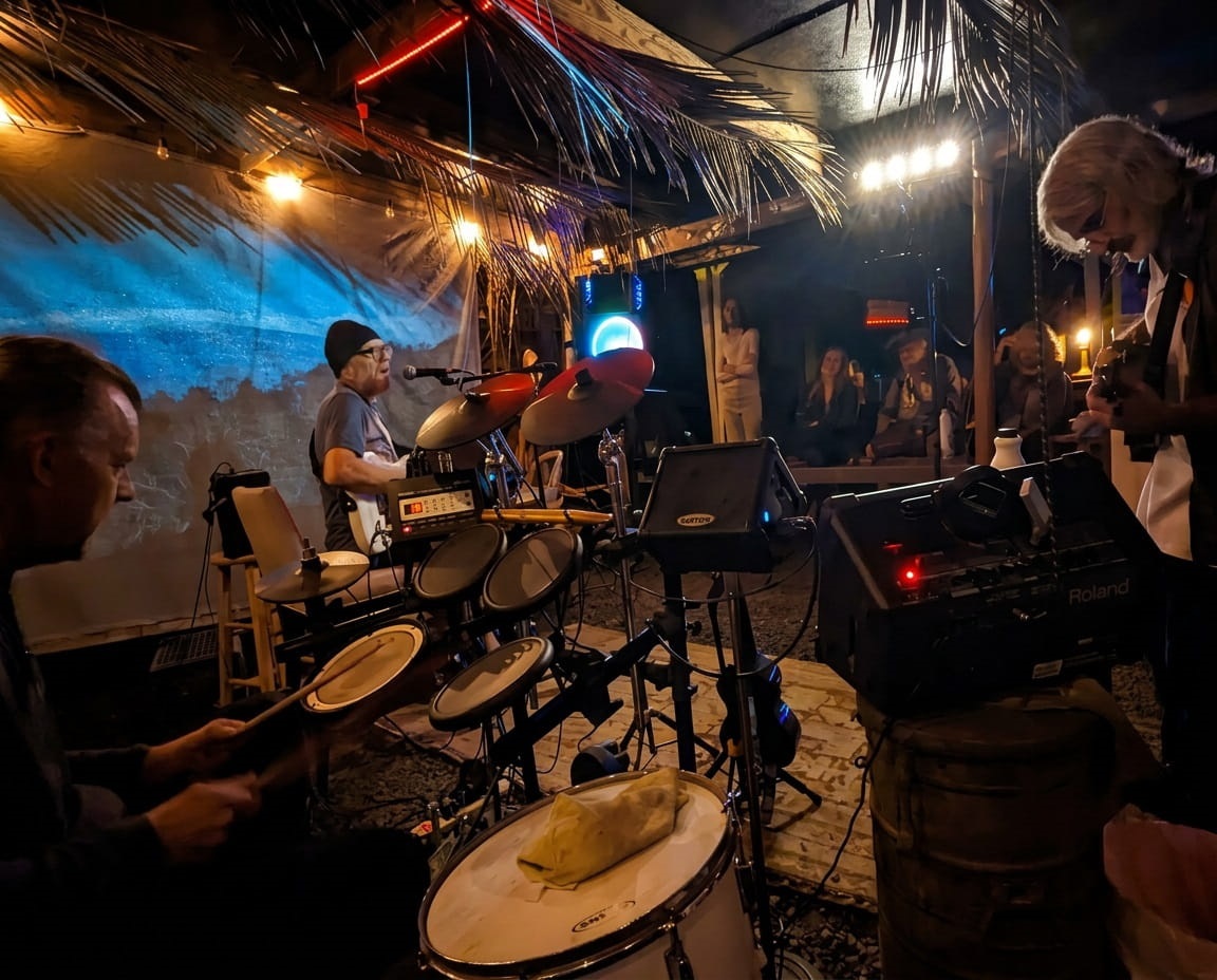 Live music performance at an open-air tiki bar at night, with drums, guitar, neon lights, and a seated crowd. Rock the Acres Open Mic with friends. Hawaiian Acres Big Island