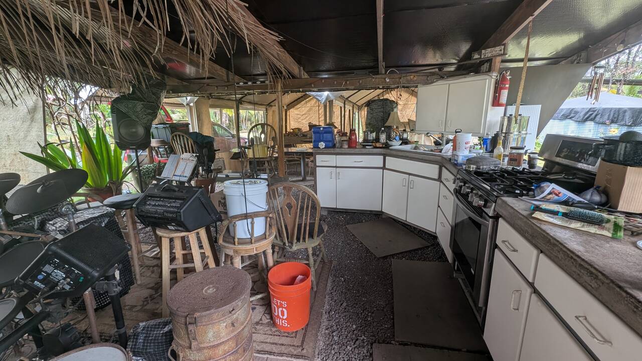 Cluttered open-air rustic kitchen with white cabinets, gas stove, and drum kit under a tropical hut roof.
